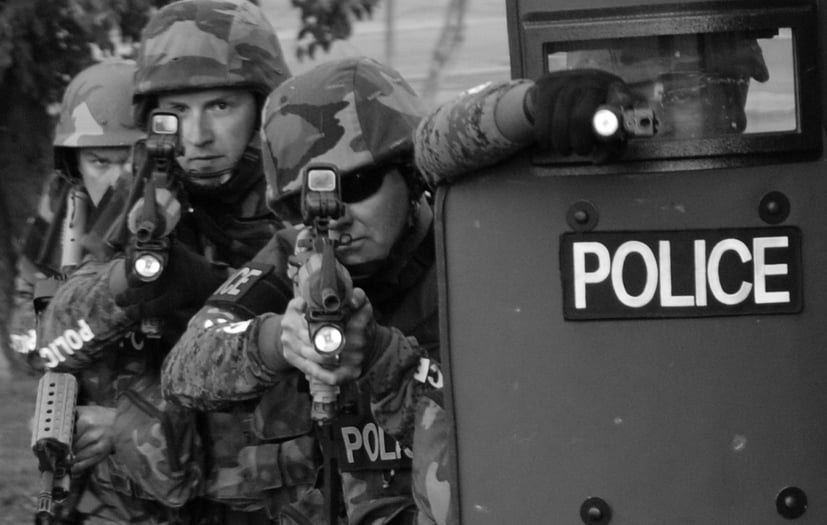 two male police officers standing behind a shield with guns aimed 