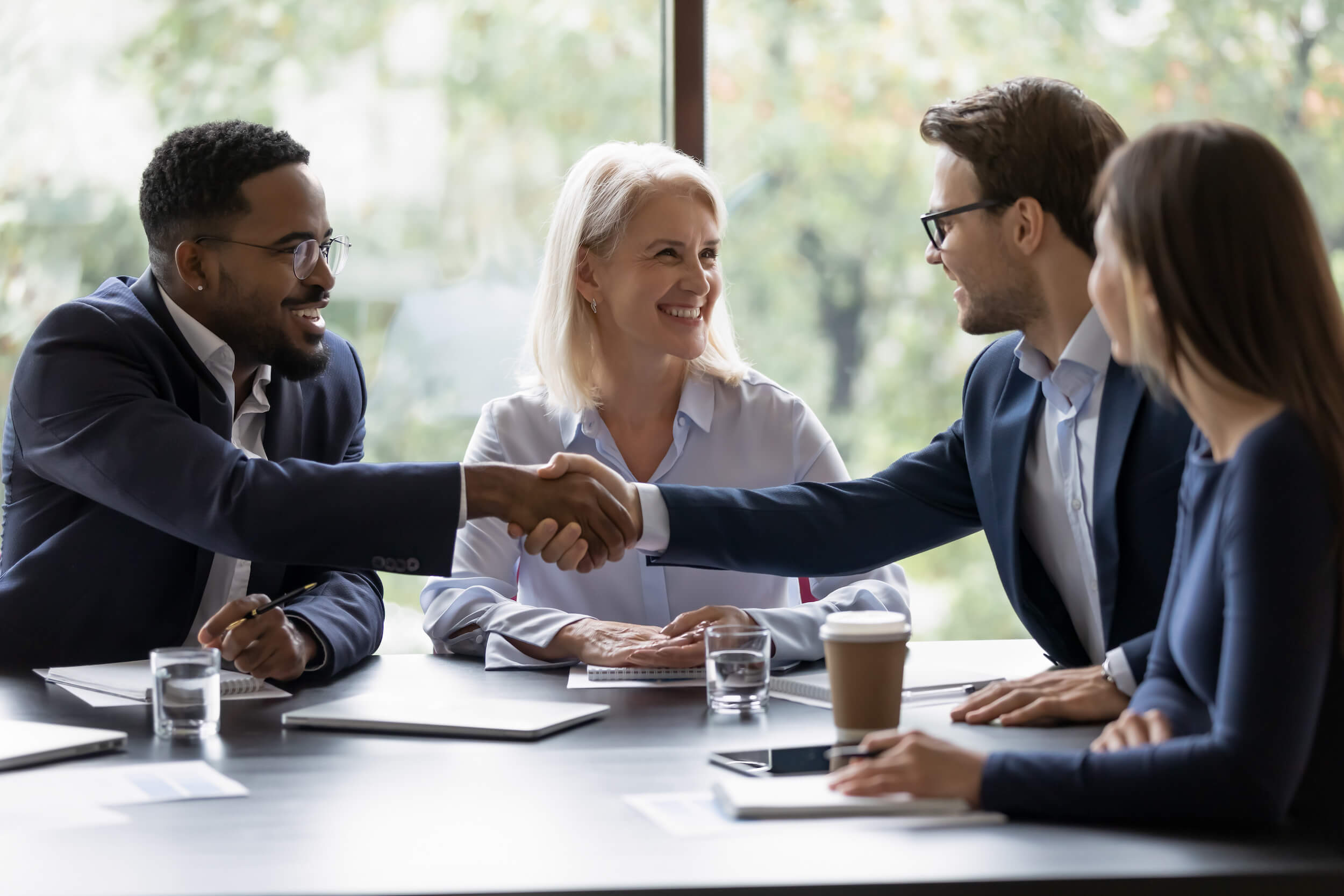 two men shake hands during a negotiation as counterparts look on